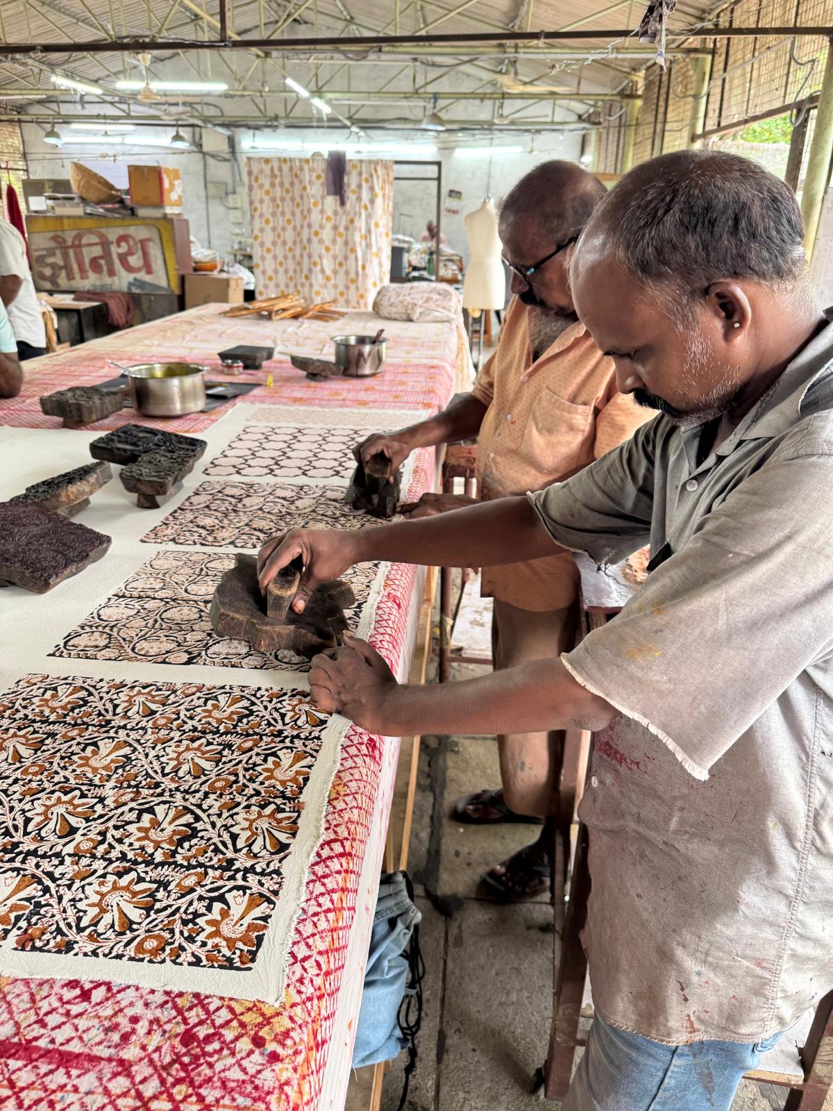 Two men working at a table in a workshop, carving and painting traditional wooden blocks with intricate floral patterns. The table is covered with carved wood pieces, and the background shows a workshop environment with other materials.