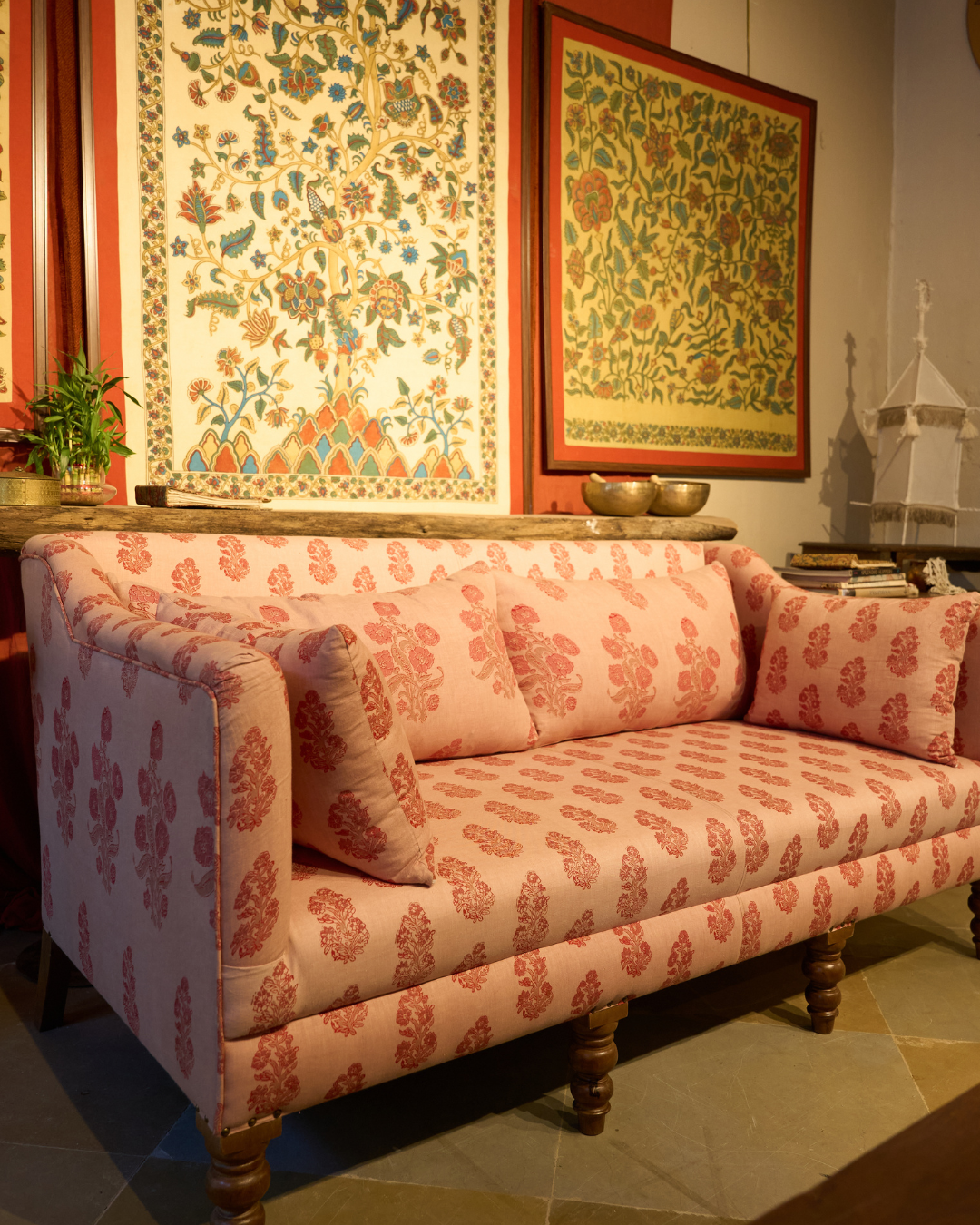 A pink upholstered sofa with a coral-red floral block print pattern, styled in a warm, traditional room setting with patterned wall hangings and wooden floor tiles.