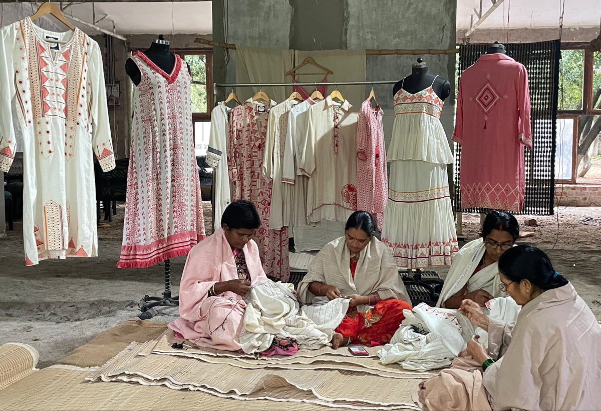 Several women sit on woven mats on the floor of a workshop, sewing and folding white and pink embroidered garments. Mannequins and clothing racks display more embroidered dresses and tunic-style pieces in the background.