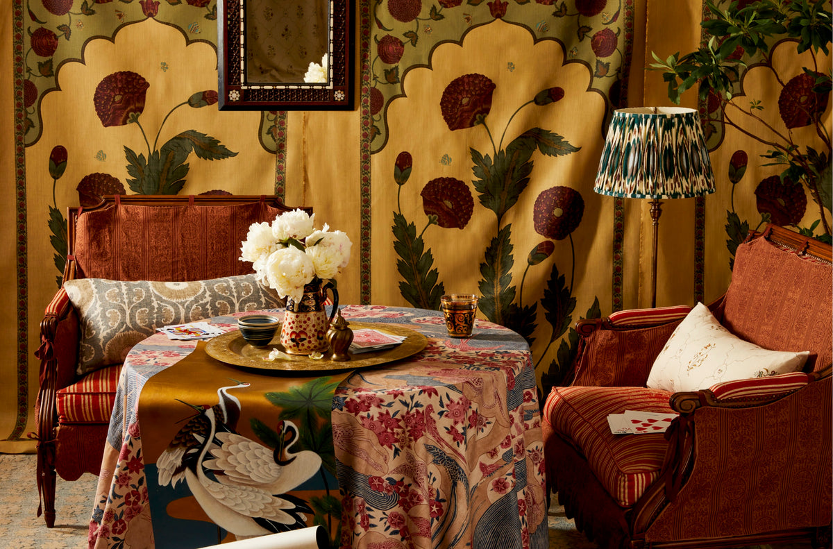A richly decorated interior scene with ornate floral wallpaper, two rust-colored armchairs, a round table covered in a detailed patterned textile, a vase of white flowers, and a patterned lamp.