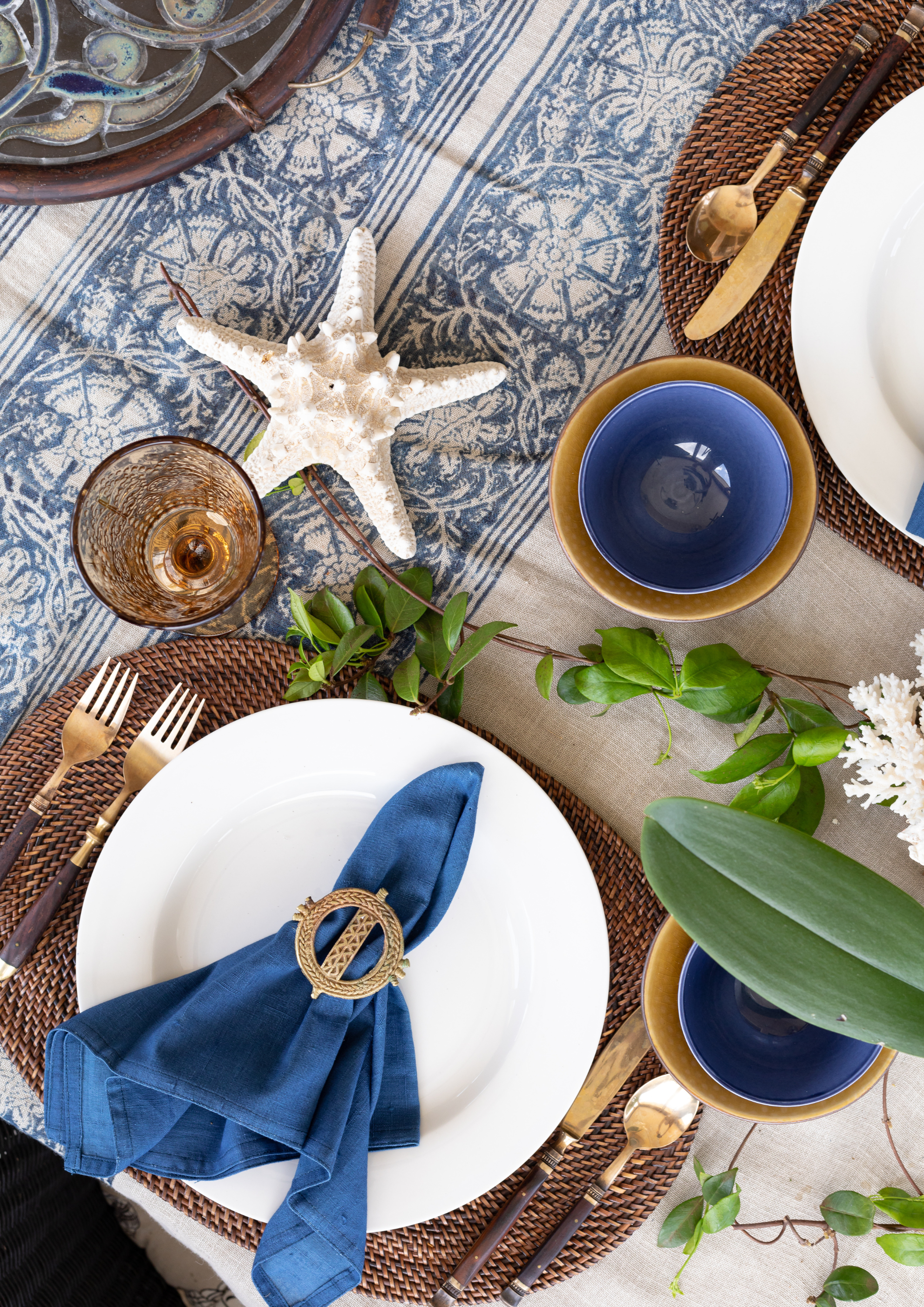 A styled nautical-themed table setting featuring a blue and white patterned table runner, woven brown placemats, white plates, deep blue bowls, gold cutlery, a blue napkin with a gold ring holder, green leaves, and a white starfish decoration.